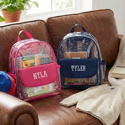 Red To Purple personalized preschool backpacks in clear PVC with a solid color flap pocket and trim, resting on a dark brown leather couch.
