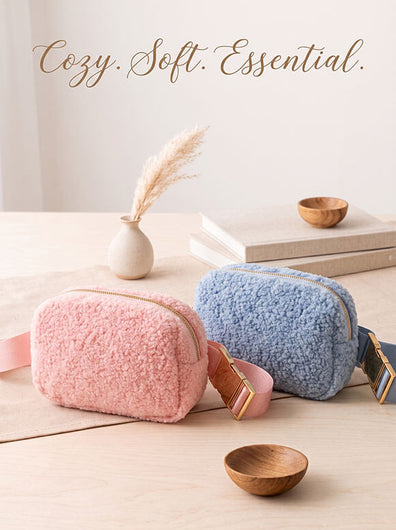 Two fuzzy zippered cosmetic pouches in soft pink and light blue are arranged on a light wood tabletop.