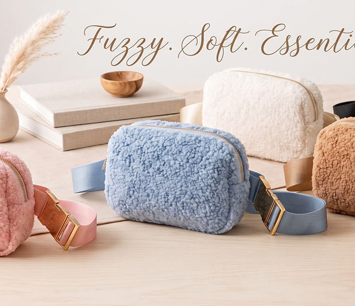 Five fuzzy fabric crossbody pouches in pink, blue, cream, camel and black, arranged on a light wooden table.